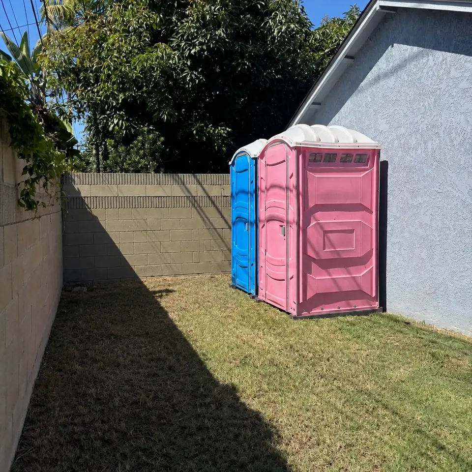Two porta potties placed on the grass in a fenced backyard